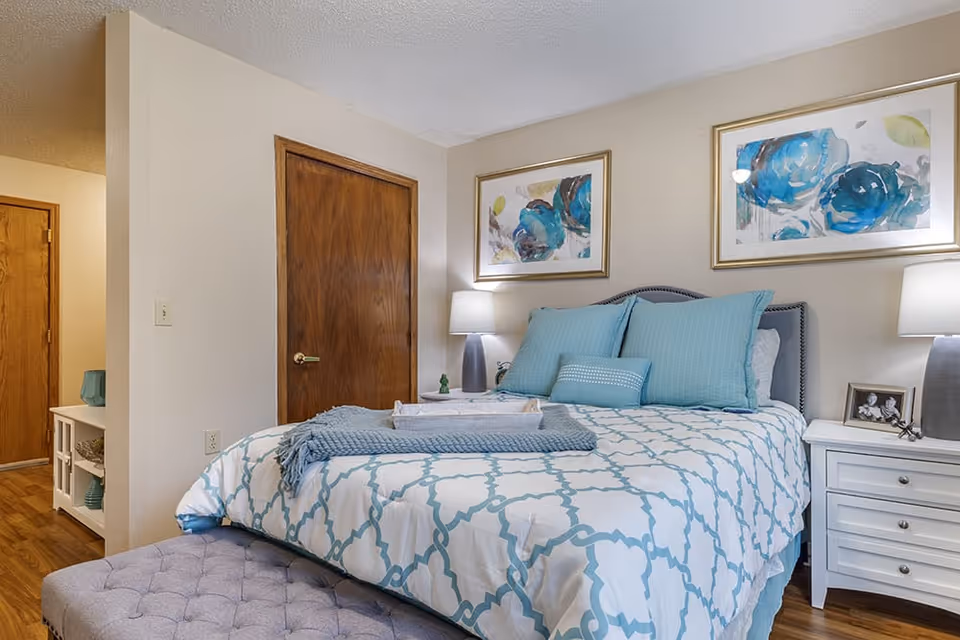 A cozy bedroom with a bed dressed in white and blue patterned bedding, multiple blue pillows, and a folded blue throw blanket. There are two framed floral paintings with blue flowers hanging above the bed. On either side of the bed are white nightstands with lamps and decorative items. The room has wooden doors and hardwood flooring.