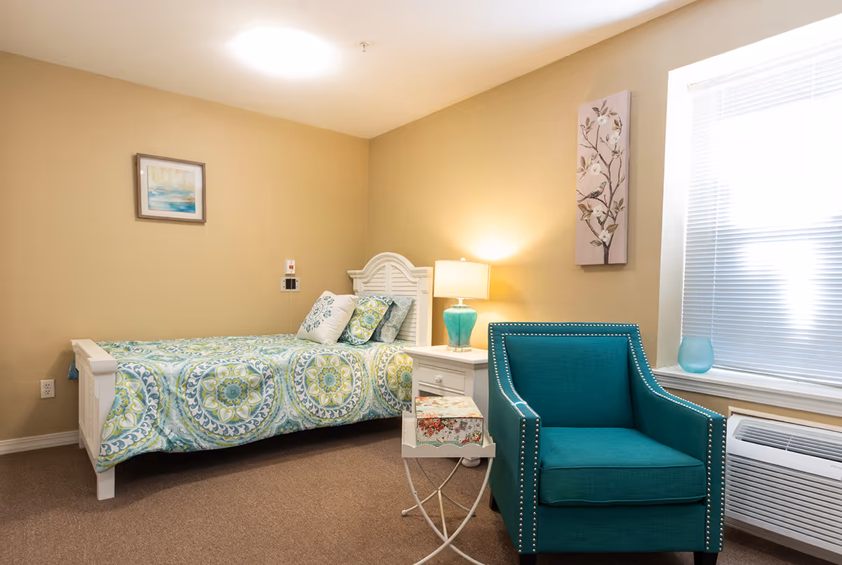 A cozy senior living bedroom with a single bed featuring a patterned blue and green bedspread and pillows. Next to the bed is a white nightstand with a turquoise lamp. A teal armchair with silver nailhead trim sits near a window with blinds. The walls are beige with two pieces of floral artwork hanging.