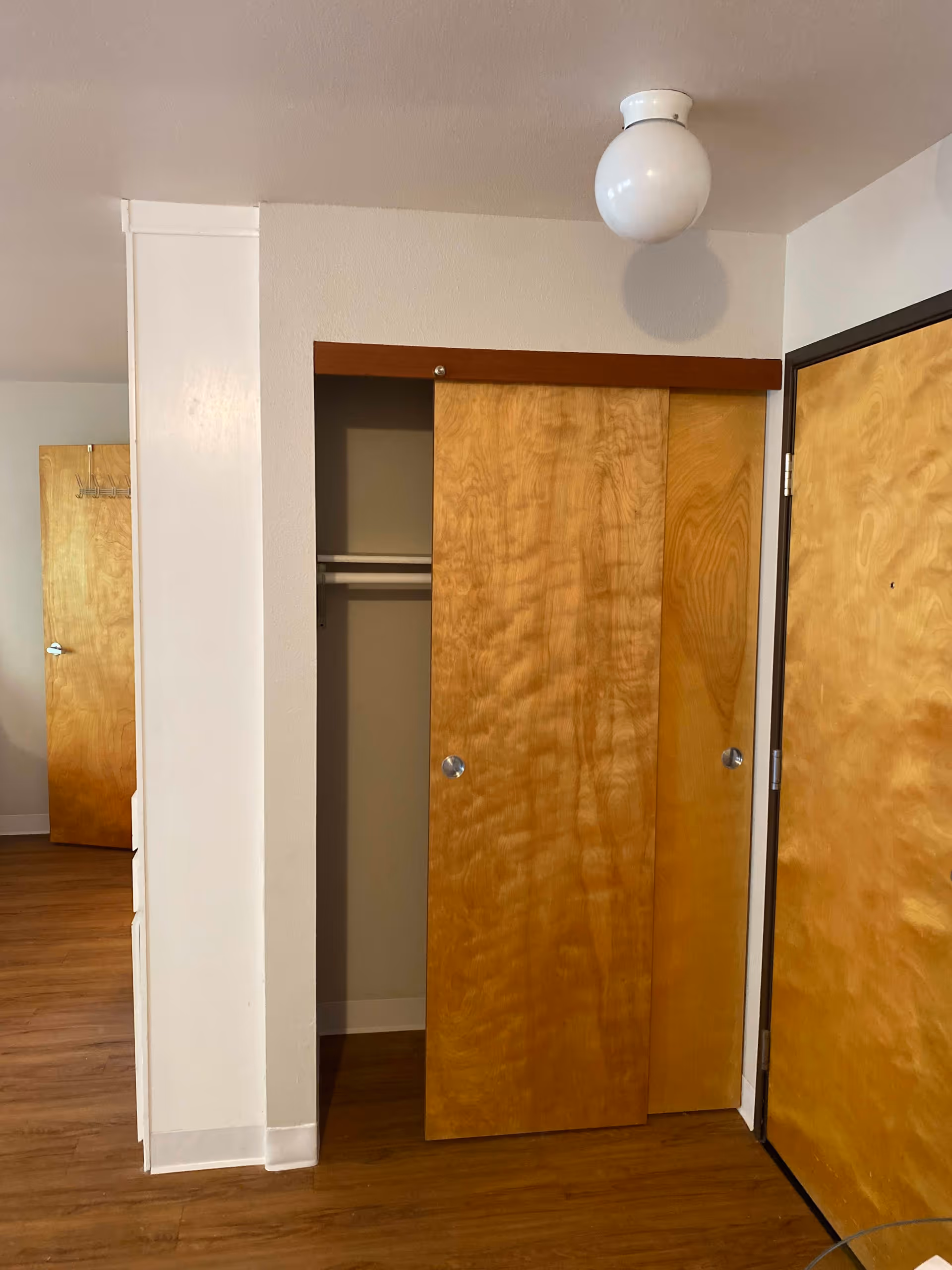 Interior view of a room showing a wooden sliding closet door partially open, a closed wooden door to the right, and another wooden door in the background. The floor is wood, and the walls are painted white. A round white ceiling light fixture is visible above the closet.