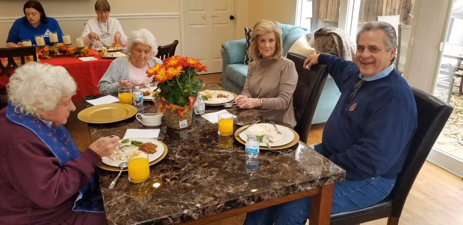 Five elderly individuals sitting around a dining table enjoying a meal together. The table has a dark marble surface with plates of food, glasses of orange juice, and a centerpiece with orange and yellow flowers. The room is well-lit with natural light coming through glass doors in the background.