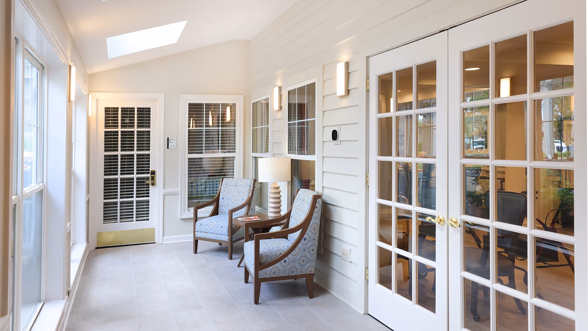 A bright and airy interior space with white walls and large windows. Two patterned armchairs with wooden frames are placed next to a small table with a lamp. There are French doors with glass panes on the right side, leading to another room with visible chairs. The floor is tiled, and the ceiling has a skylight allowing natural light to fill the room.