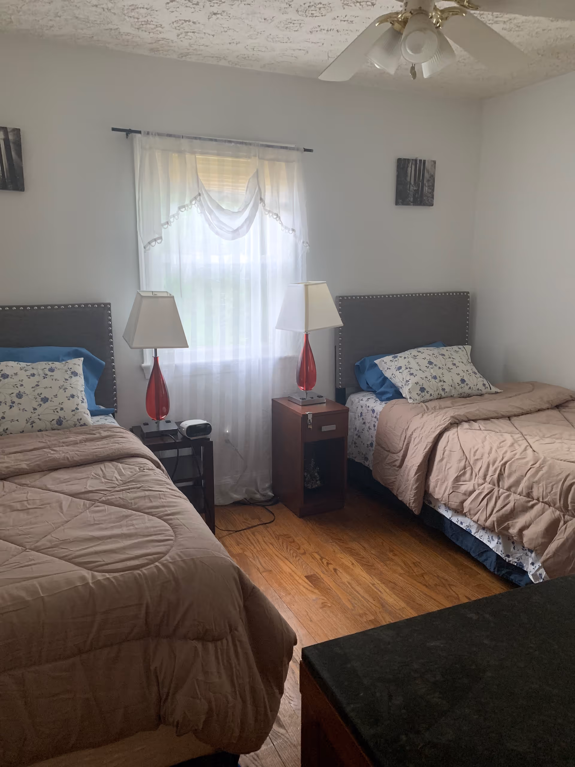 A bedroom with two twin beds, each with a beige comforter and floral pillows. Between the beds is a small wooden nightstand with a red lamp and a clock. A window with sheer white curtains is centered on the wall behind the nightstand. The room has hardwood floors and a ceiling fan with lights.