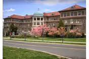Large multi-story stone building with a green dome on the roof, surrounded by trees including a pink flowering tree, with a road and grassy area in the foreground under a partly cloudy sky.