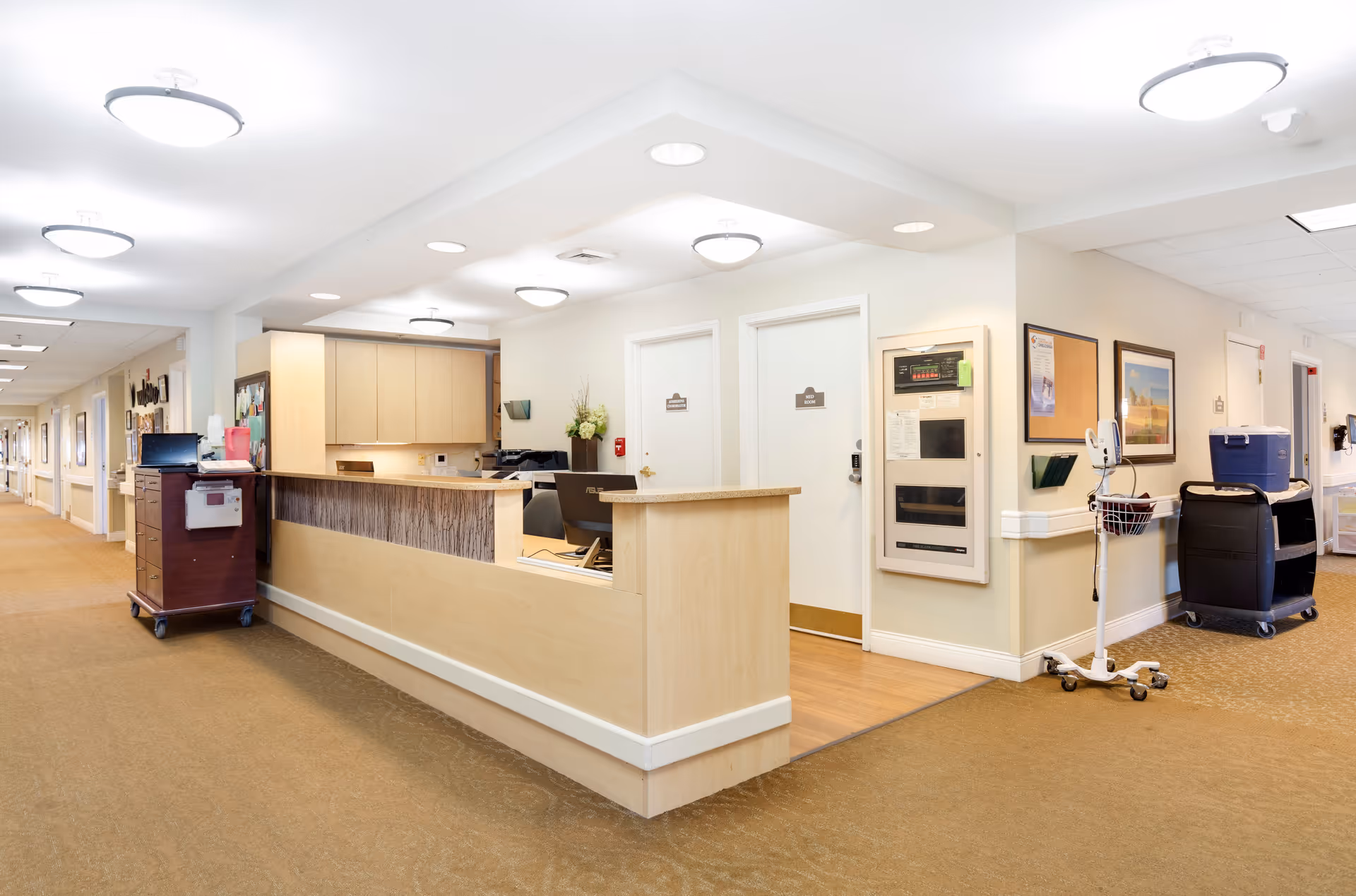 Reception desk area in a senior living facility hallway with beige walls, overhead lights, and medical carts. Two white doors are visible behind the desk, along with framed pictures and a bulletin board on the wall.
