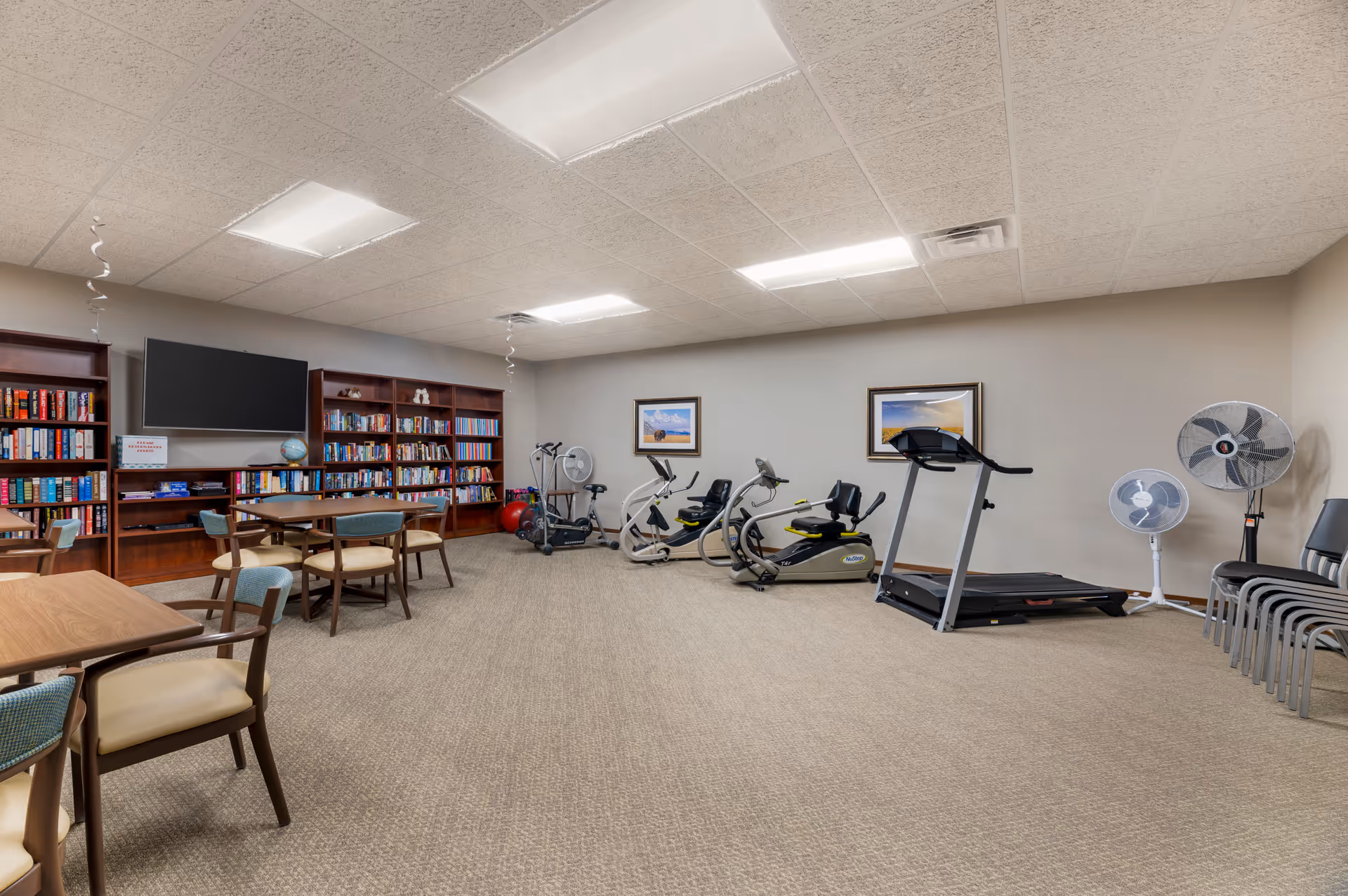 A spacious senior living facility room with exercise equipment including stationary bikes and a treadmill on the right side. On the left side, there are tables and chairs arranged for seating, with bookshelves filled with books and a wall-mounted TV above them. The room has beige walls, carpeted floor, and ceiling lights.