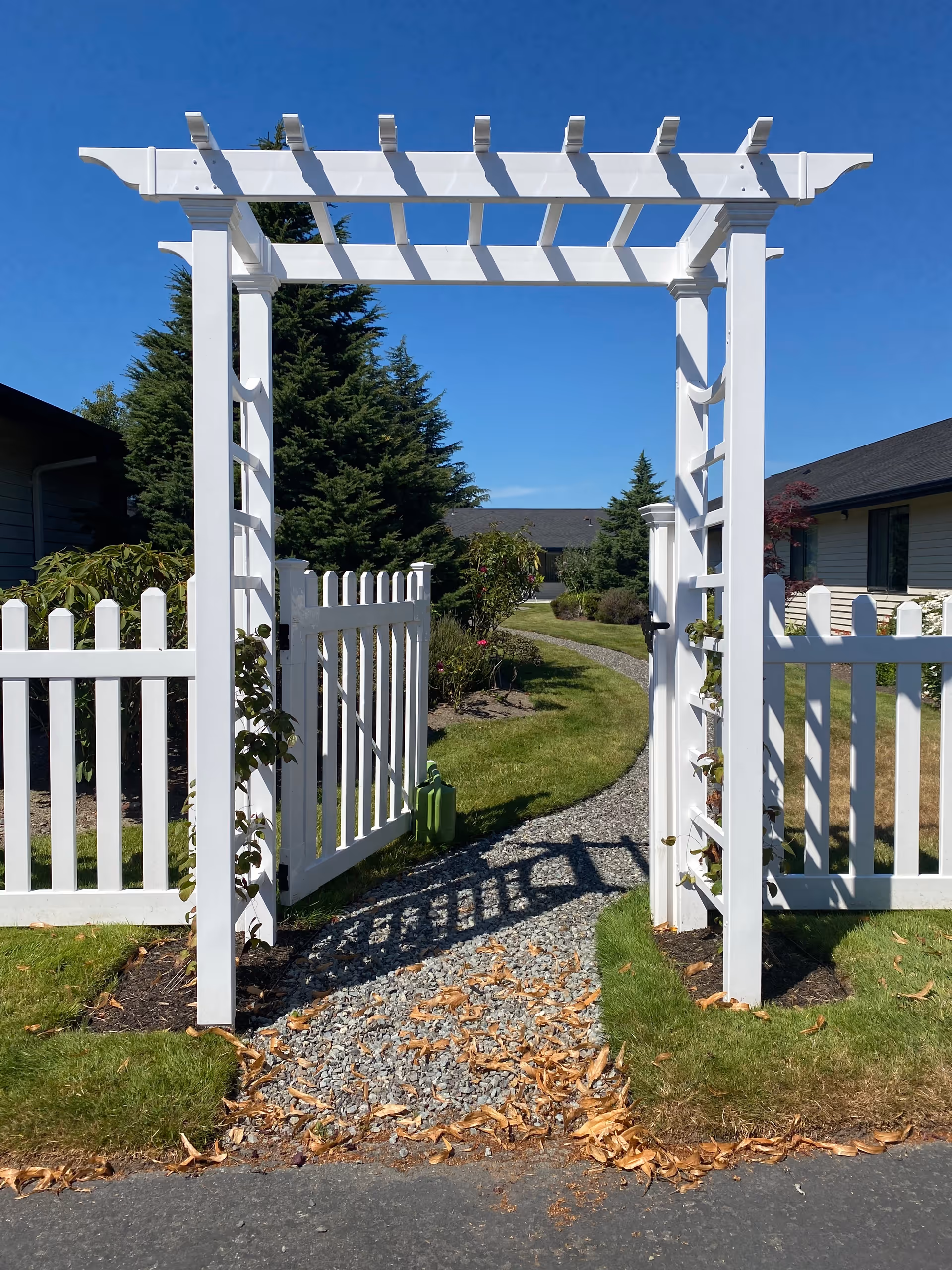 White wooden garden archway with a white picket fence gate open, leading to a gravel pathway surrounded by green grass and shrubs under a clear blue sky.