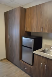 Interior view of a small kitchenette area featuring a compact stainless steel refrigerator, wooden cabinets above and beside it, a countertop with a built-in sink, and wood-look flooring.