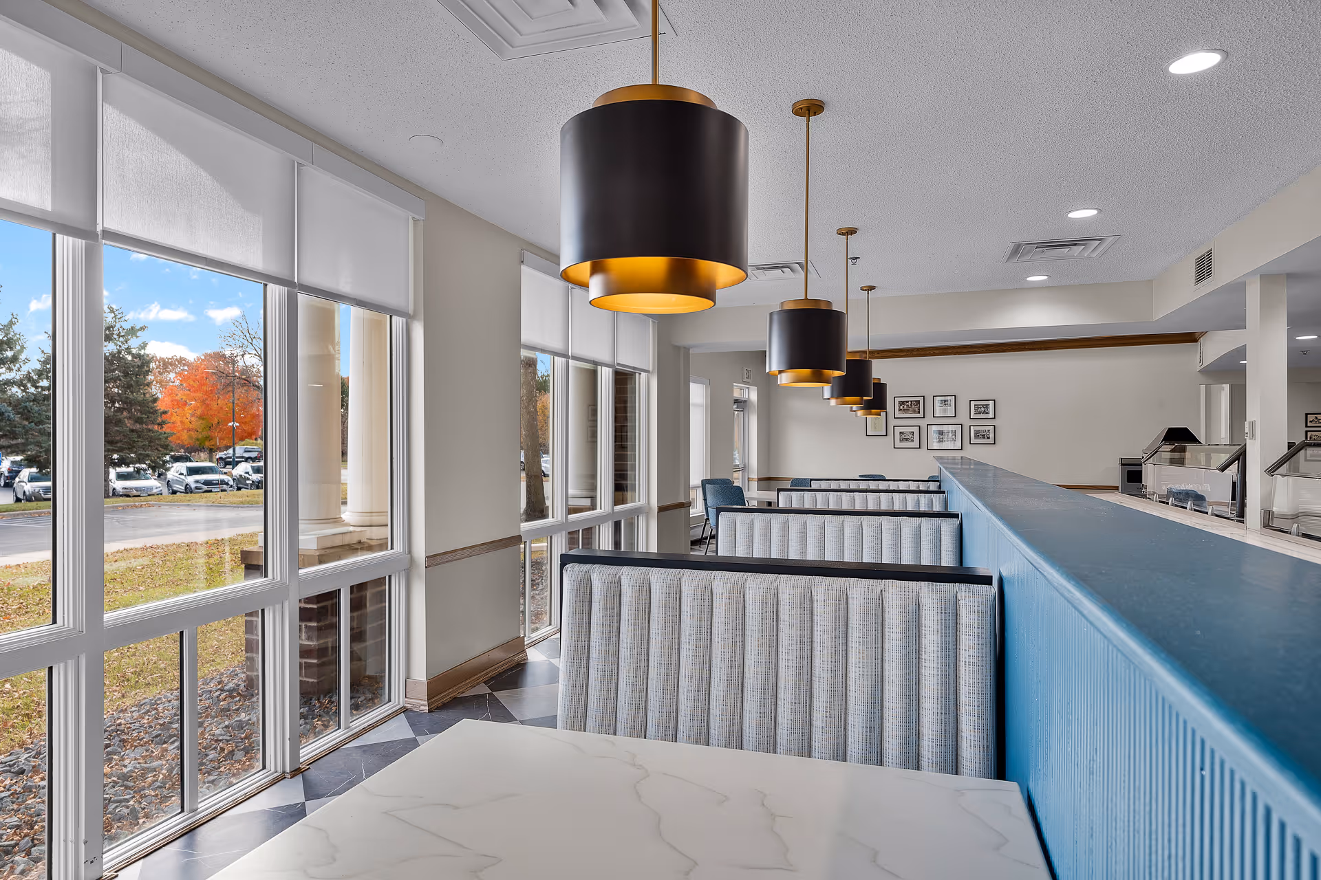 Interior view of a dining area with booth seating and large windows showing an outdoor parking lot with trees. The booths have light gray upholstery and white marble-like tabletops. Modern black and gold pendant lights hang from the ceiling above the booths. The walls are light-colored with framed pictures and the floor has a dark tile pattern.