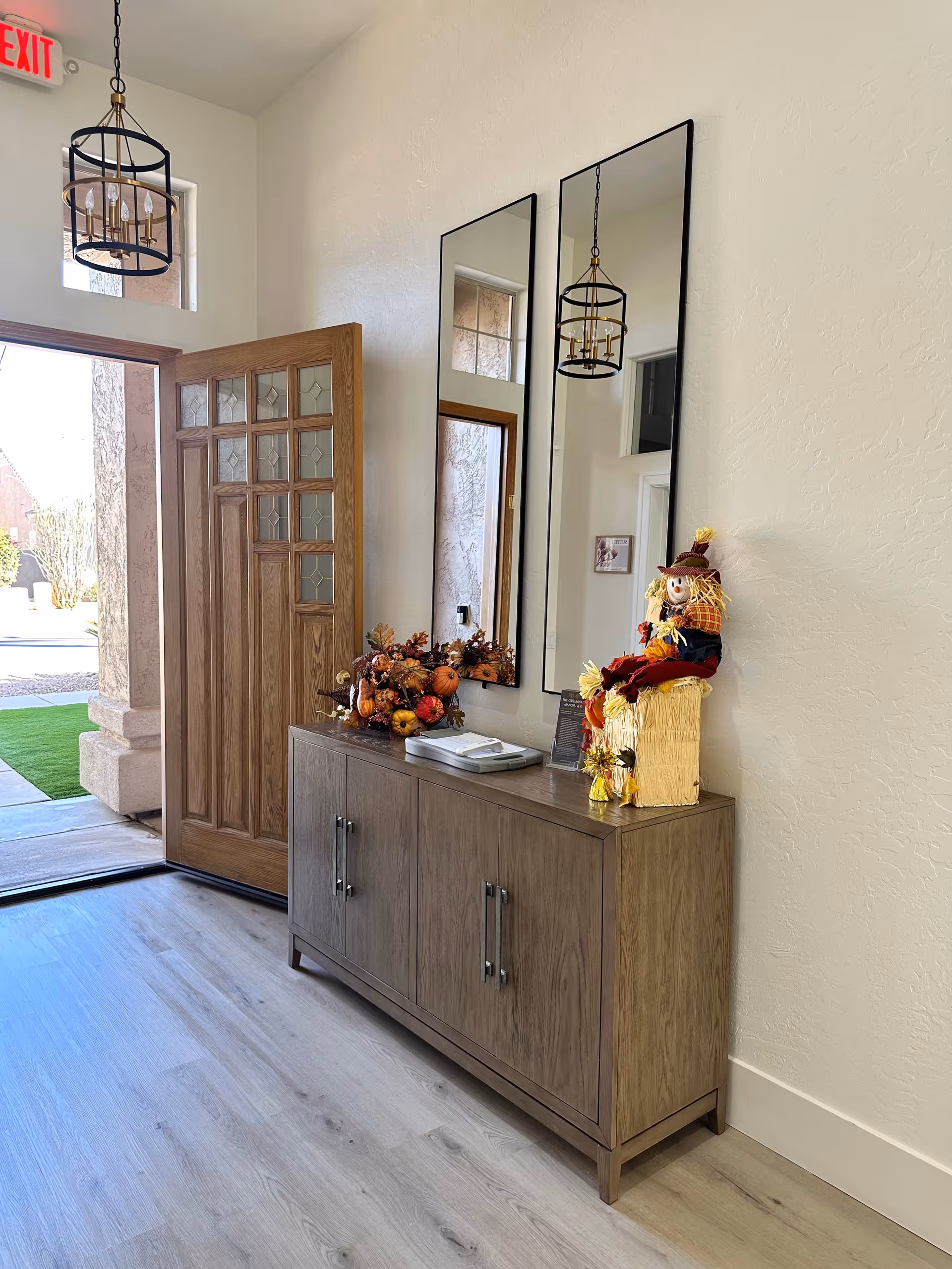 Open entryway with a wooden front door, a console table with fall decorations and a scarecrow, and two tall mirrors on a white wall.