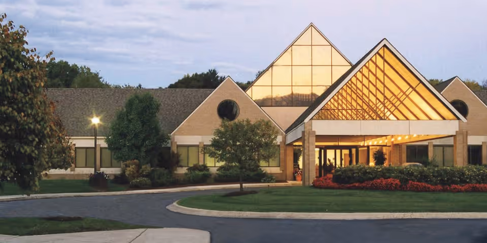 Exterior view of Shelby - An Optalis Health & Rehabilitation Center building during dusk, featuring a triangular glass entrance with warm interior lighting, surrounded by trees, shrubs, and a curved driveway.