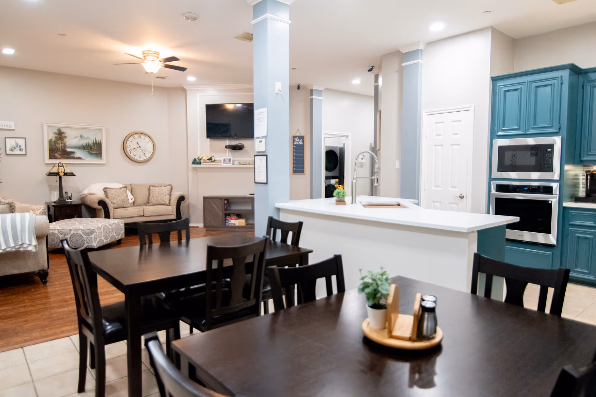 Interior view of a senior living facility showing a combined dining and living area. In the foreground, there are dark wooden dining tables and chairs with a small plant and condiments on one table. The background features a cozy living room with beige sofas, a patterned ottoman, a wall clock, and a TV mounted above a fireplace. To the right, there is a kitchen area with teal cabinets, a white countertop island with a sink, and built-in microwave and oven.