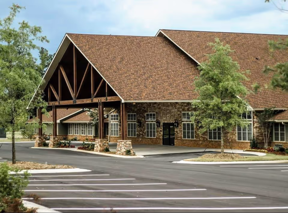 Exterior view of The Timberley facility showing a large building with a brown shingled roof, stone and brick facade, multiple windows, and a covered entrance supported by wooden beams. The surrounding area includes a paved parking lot and landscaped greenery with trees and shrubs.