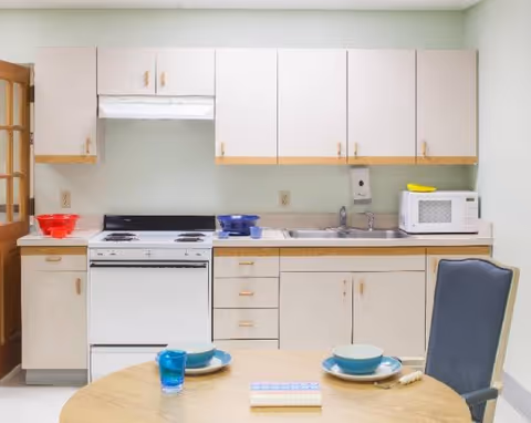 A small communal kitchen with cabinets, a stove, sink, microwave, and a round dining table.