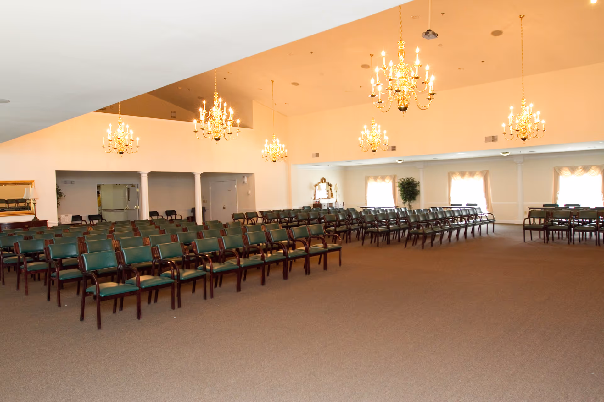 A large room with rows of green cushioned chairs arranged facing forward. The room has beige walls, several chandeliers hanging from the ceiling, and windows with curtains letting in natural light. There is a mirror and a potted plant along the far wall.