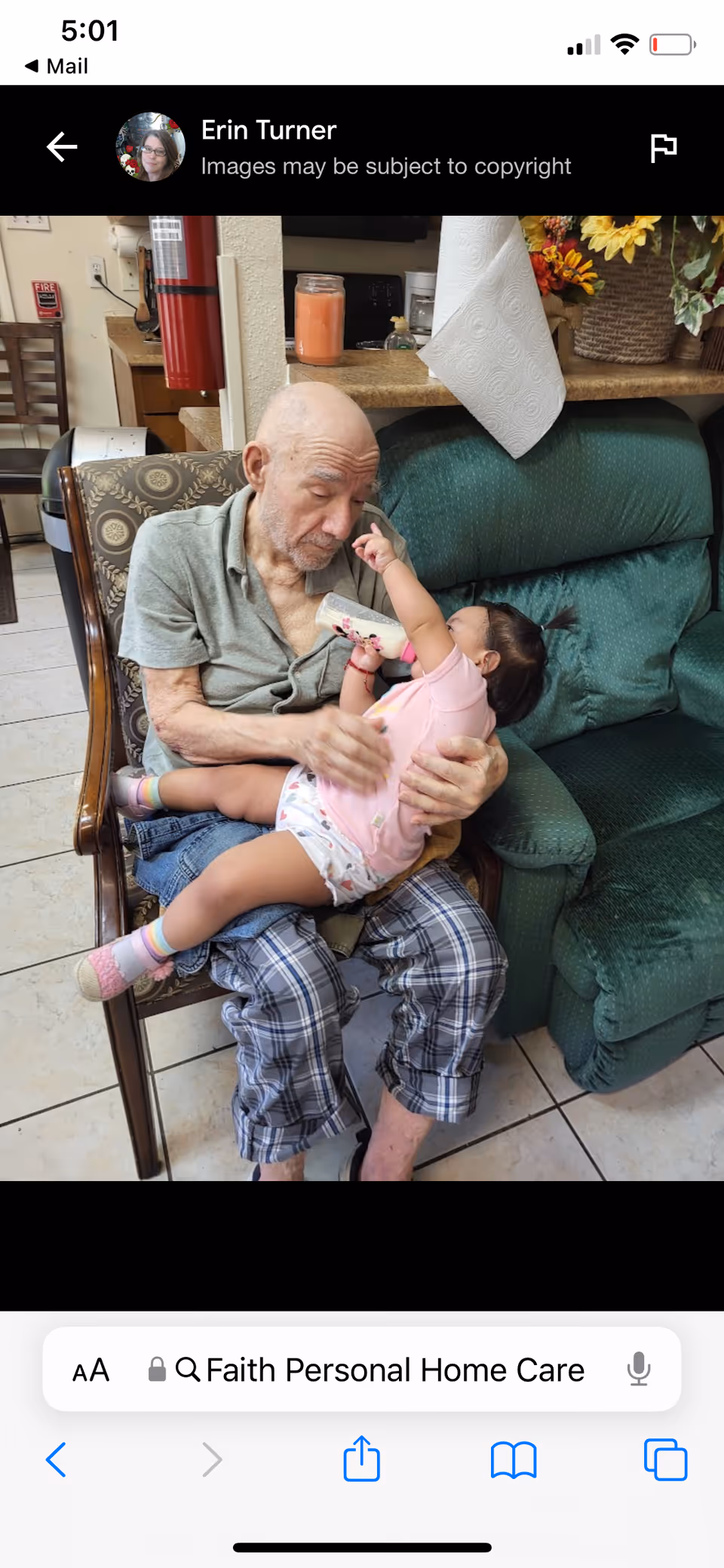 An elderly man sitting on a chair indoors, holding and feeding a baby with a bottle. The setting includes a green cushioned chair, a table with a paper towel roll, a candle, and a basket with flowers in the background.