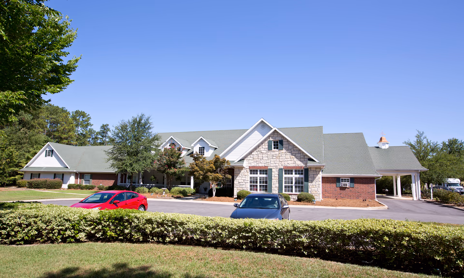 Front exterior of a single-story senior living building with stone and brick facade, green roof, driveway and parked cars.