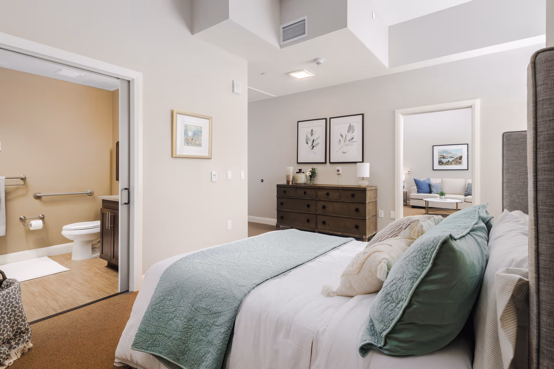 A bright and cozy bedroom with a neatly made bed featuring white and green bedding. The room has beige walls and carpeted floors. To the left, there is an open doorway leading to a bathroom with a toilet, grab bars, and a towel rack. In front of the bed, there is a dark wooden dresser with decorative items and two framed botanical prints on the wall above it. Through another open doorway, a living room area with a sofa, cushions, and a framed picture on the wall is visible.