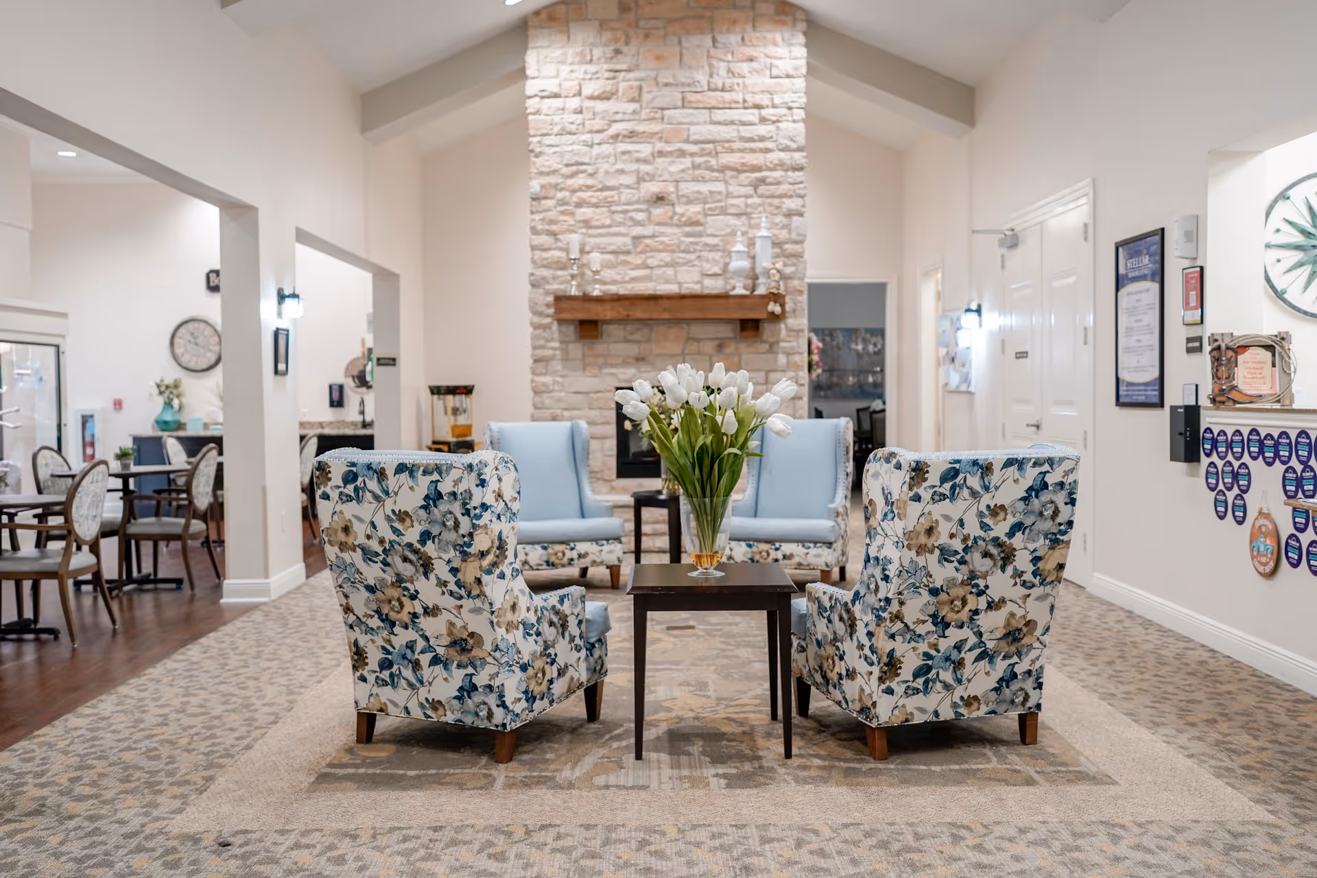 A bright senior living lounge with floral armchairs, a small table holding white tulips, light blue seating and a stone fireplace in the background.