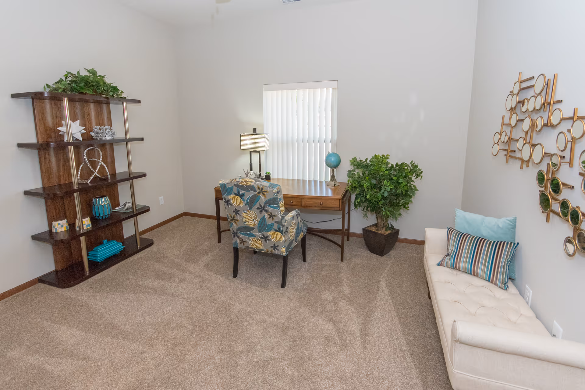 Furnished living room with a wooden desk and patterned chair centered under a window, shelving on the left, and a tufted bench with decorative pillows on the right.