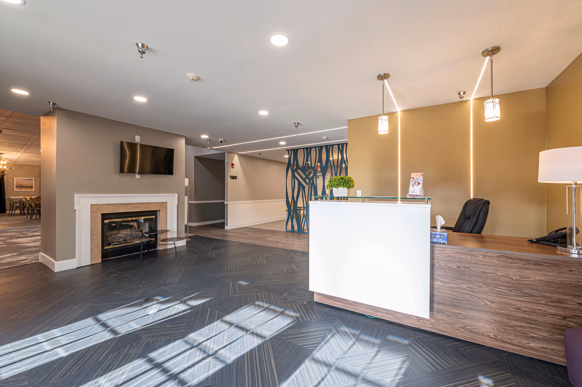 Bright reception lobby with a front desk, fireplace and mounted TV in a senior living facility.