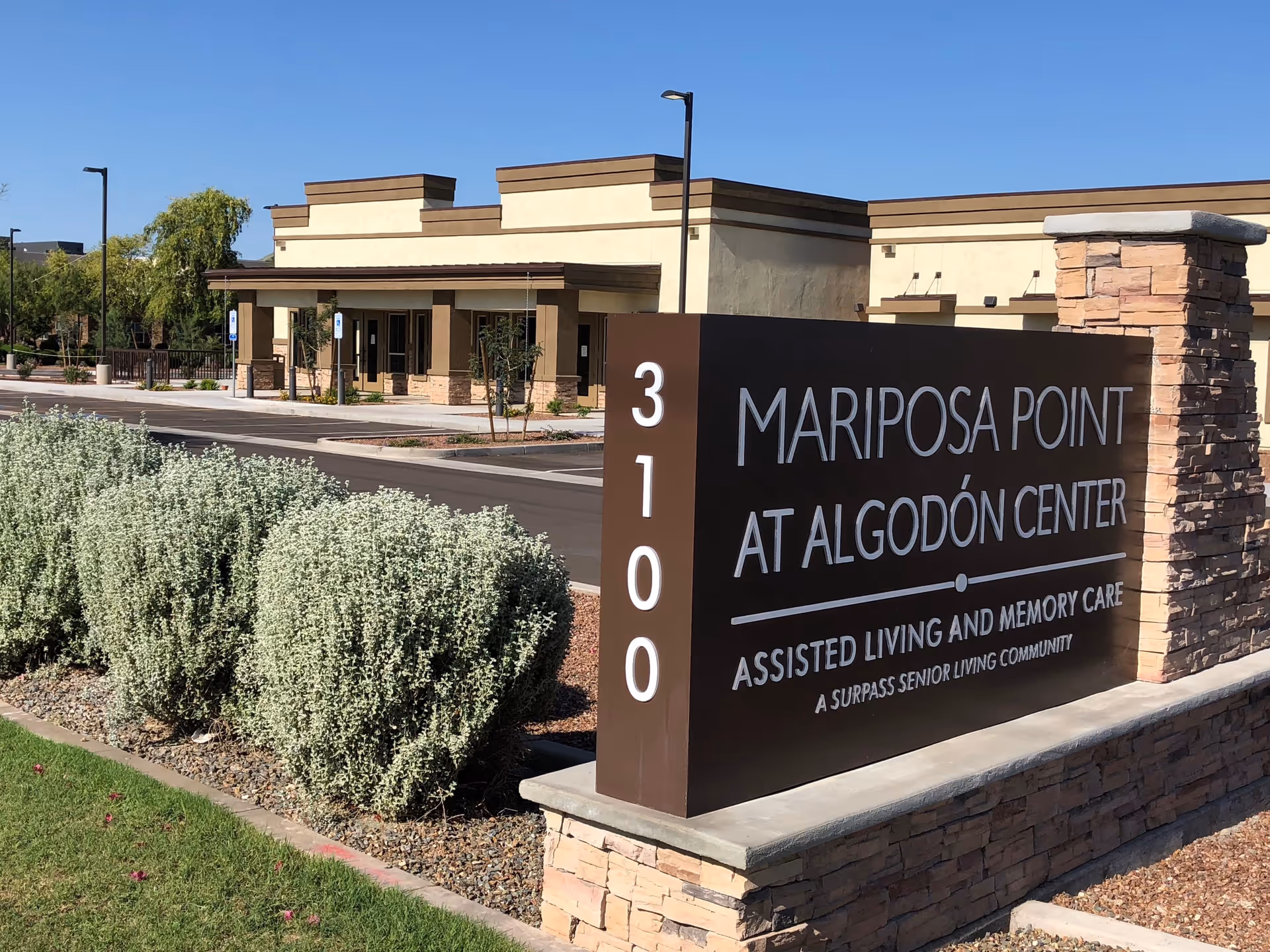 Exterior view of Mariposa Point at Algodón Center, an assisted living and memory care facility. The image shows a large brown sign with white text displaying the facility name and address number 3100. Behind the sign is a beige building with brown trim, a parking lot, and some landscaping with bushes and trees under a clear blue sky.