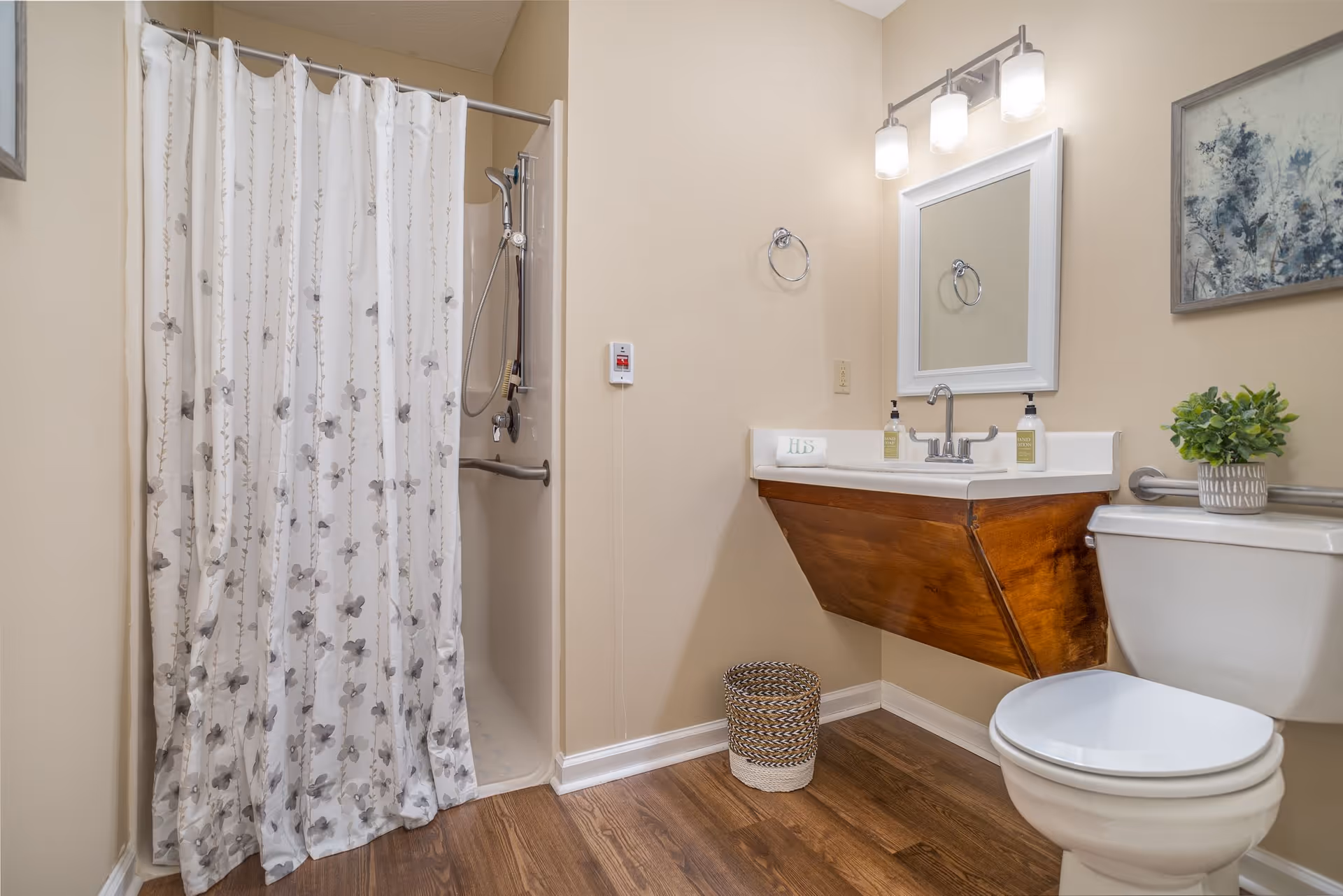 A bathroom with a shower area covered by a floral shower curtain, a wooden floating sink vanity with a mirror and two light fixtures above it, a toilet with a small potted plant on the tank, a woven wastebasket on the wooden floor, and a framed artwork on the wall.