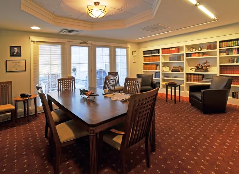 A well-lit room with a large wooden table surrounded by six chairs in the center. The room has a red patterned carpet and a ceiling light fixture. There are two black leather armchairs near built-in white bookshelves filled with books and decorative items. French doors with blinds lead to an outdoor area with a swing and a grill. Framed pictures and certificates hang on the walls.