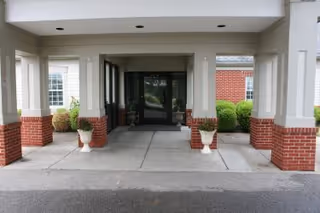 Covered entrance to a building with white columns and brick bases, two white planters with greenery on either side of the walkway, and glass double doors at the end of the path.