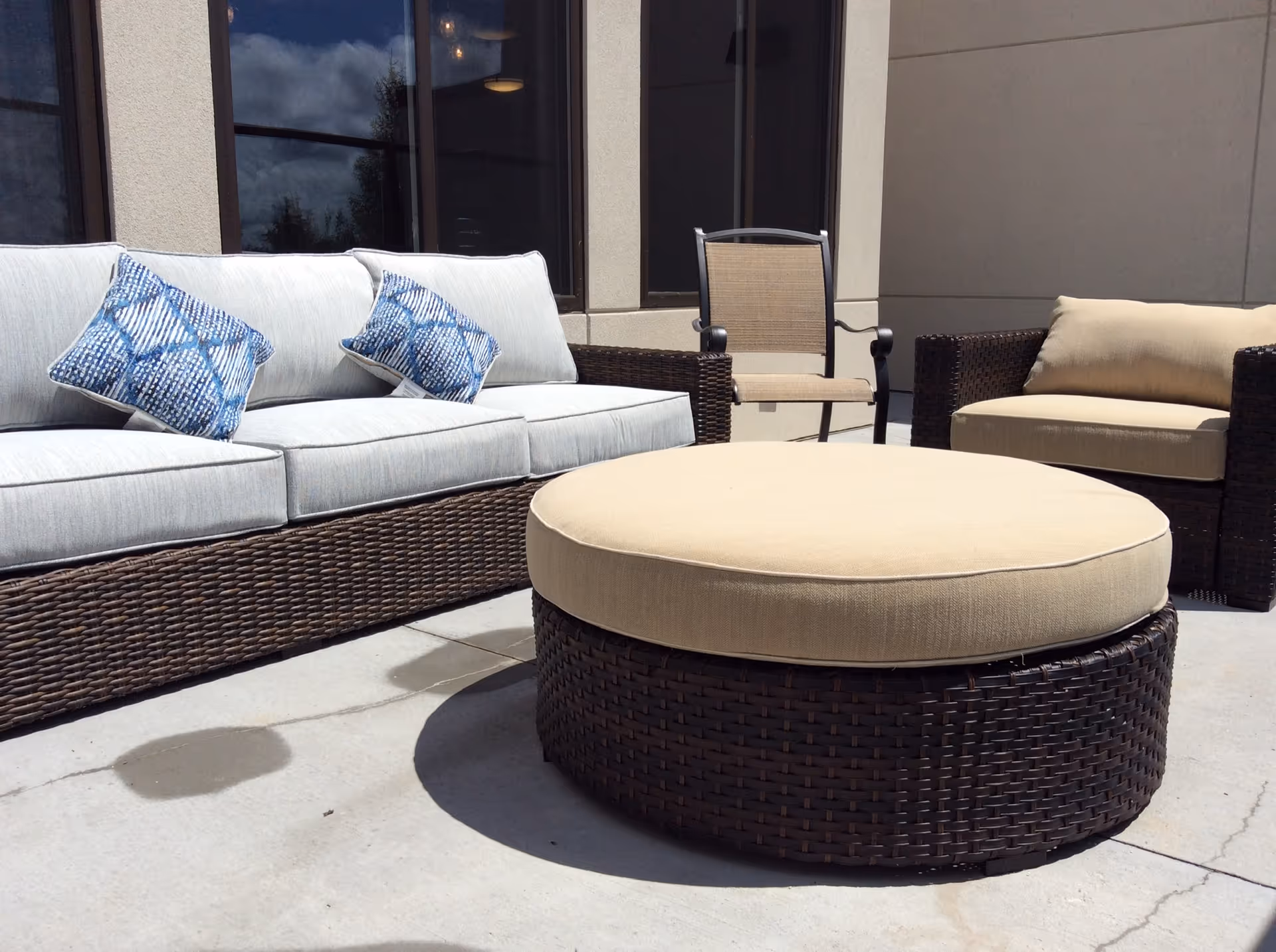 Outdoor patio area with wicker furniture including a cushioned sofa with blue patterned pillows, a cushioned armchair, a beige cushioned round ottoman, and a chair with a metal frame and beige seat. The setting is on a concrete floor with building windows and walls in the background.