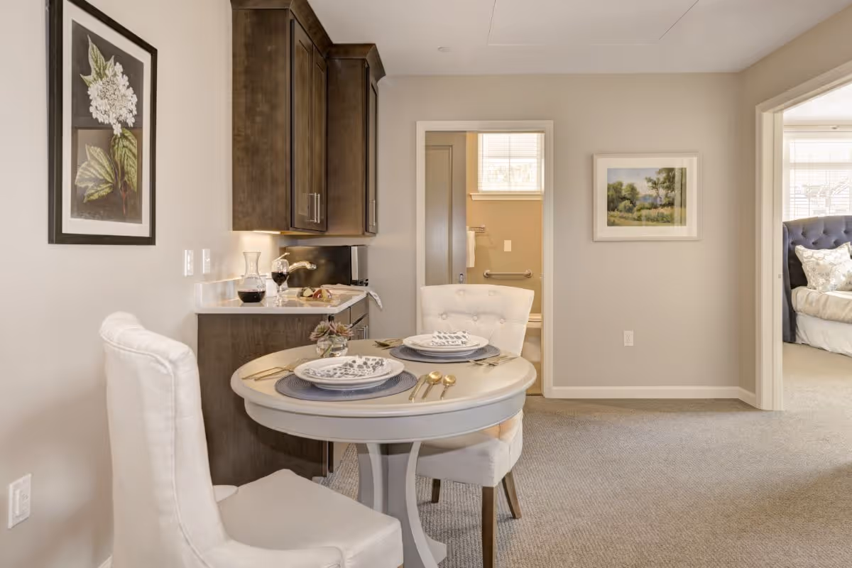 A cozy dining area with a small round table set for two with plates, napkins, and gold utensils. Two upholstered chairs are placed around the table. To the left, there is a kitchenette with dark wood cabinets, a countertop with a wine decanter and glass, and a small plate of food. The background shows a doorway leading to a bathroom with grab bars and a window, and another doorway leading to a bedroom with a bed and pillows. The walls are painted light beige and decorated with framed artwork.
