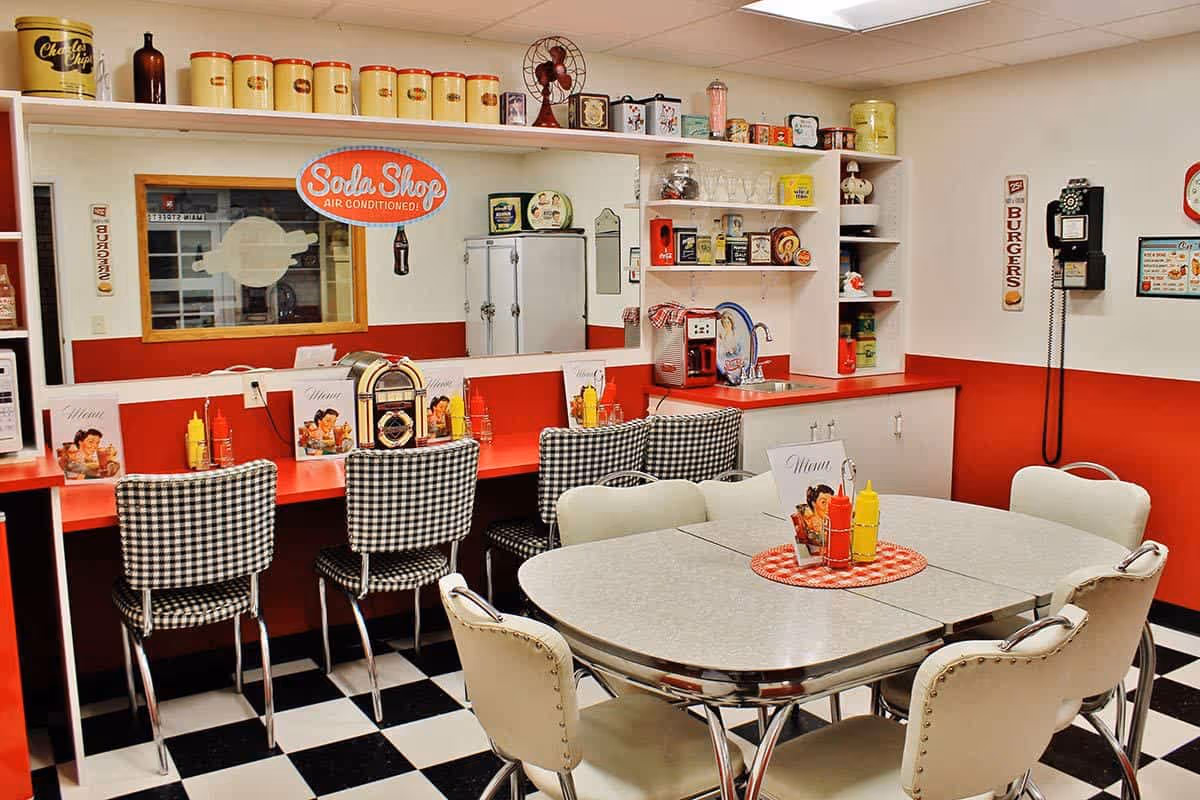 A retro-style diner interior with red and white decor, black and white checkered floor, a counter with black and white checkered chairs, a round table with white cushioned chairs, condiment bottles, menus, a vintage jukebox, a soda shop sign, and various vintage kitchen items and decorations on shelves and walls.