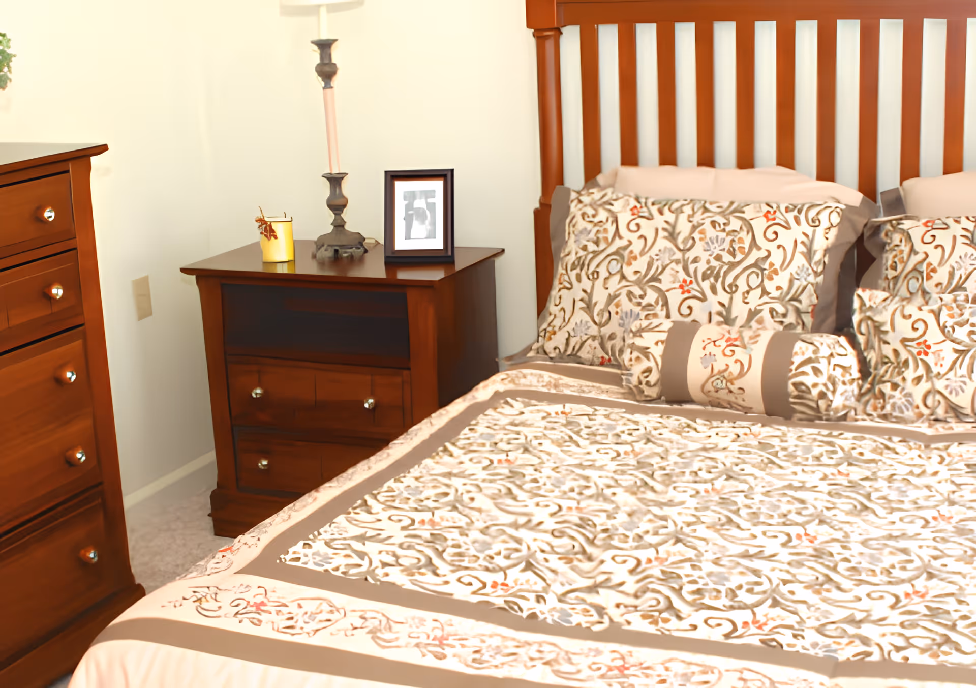 A bedroom with a wooden bed frame and a bed made with a patterned bedspread and matching pillows in beige, brown, and muted floral designs. Next to the bed is a wooden nightstand with a candle, a tall candle holder, and a framed photo. To the left is a wooden dresser with multiple drawers.