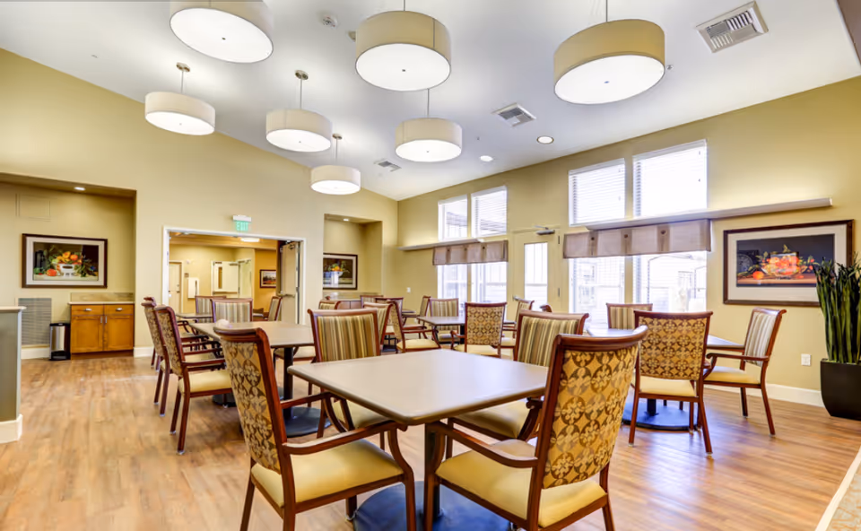A bright and spacious dining room with multiple tables and chairs arranged neatly. The room has large windows with blinds, framed artwork on the walls, and modern ceiling lights. The floor is wooden, and there is a doorway leading to another room.