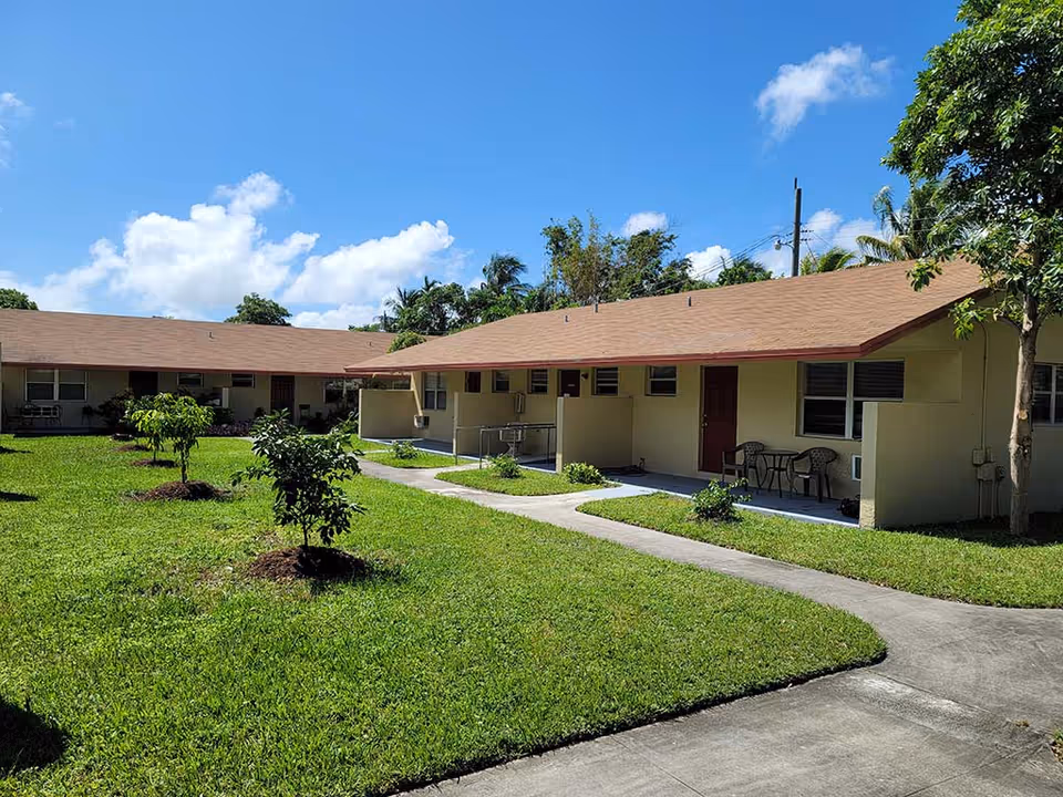 Single-story beige residential units around a grassy courtyard with a paved walkway and outdoor seating under a blue sky.
