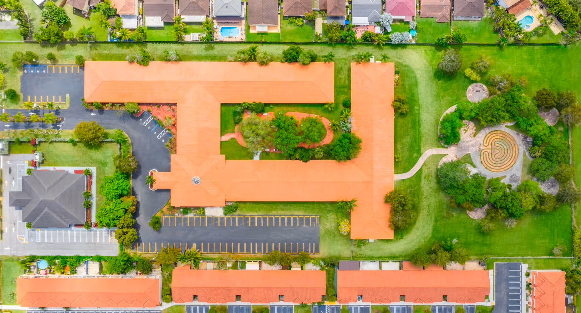 Aerial view of a senior living complex with orange-roofed buildings, parking lots, landscaped courtyards and a circular walking maze.