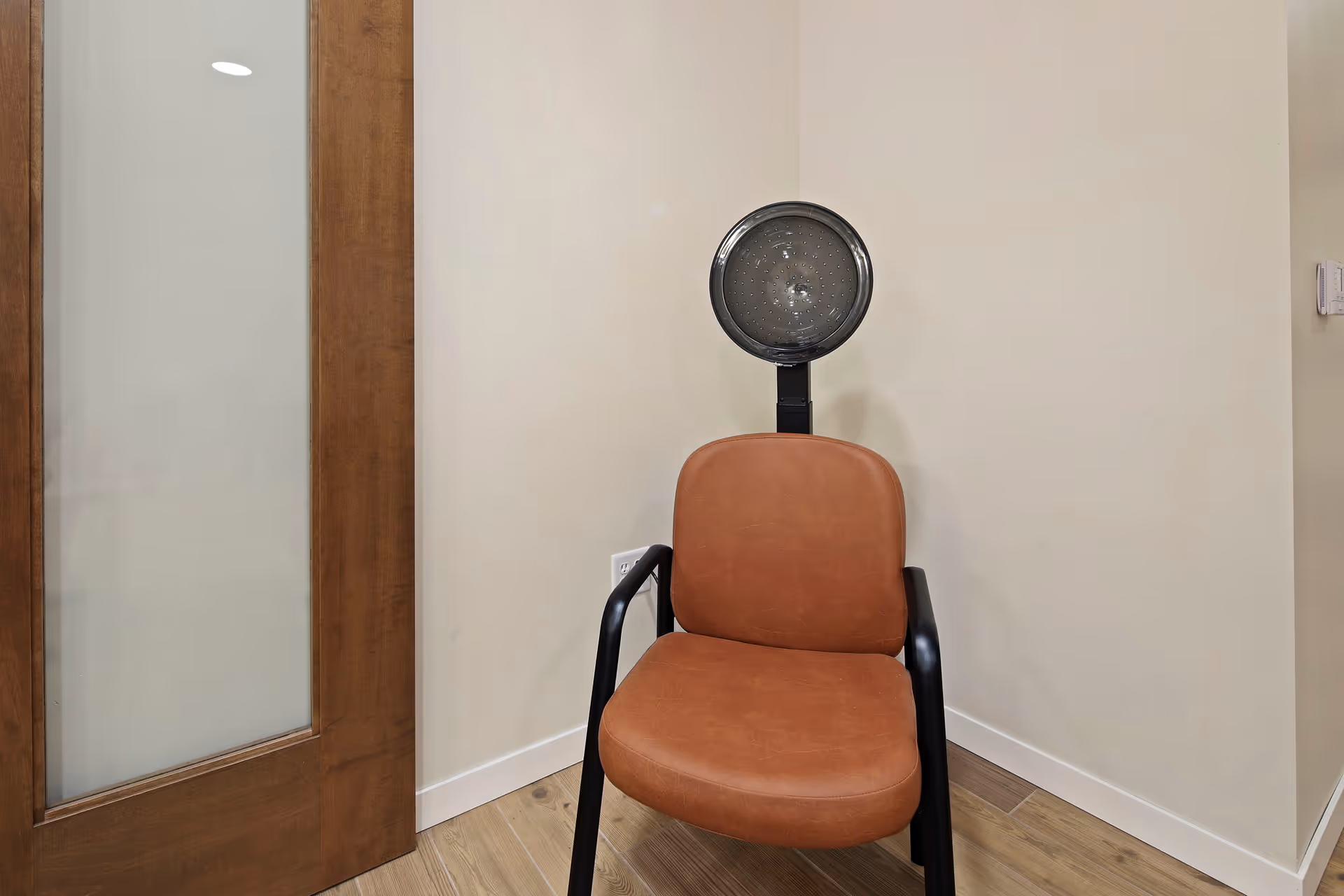 A brown leather chair with black armrests positioned against a beige wall, with a round hair dryer hood attached behind the chair, next to a wooden door with frosted glass panel.