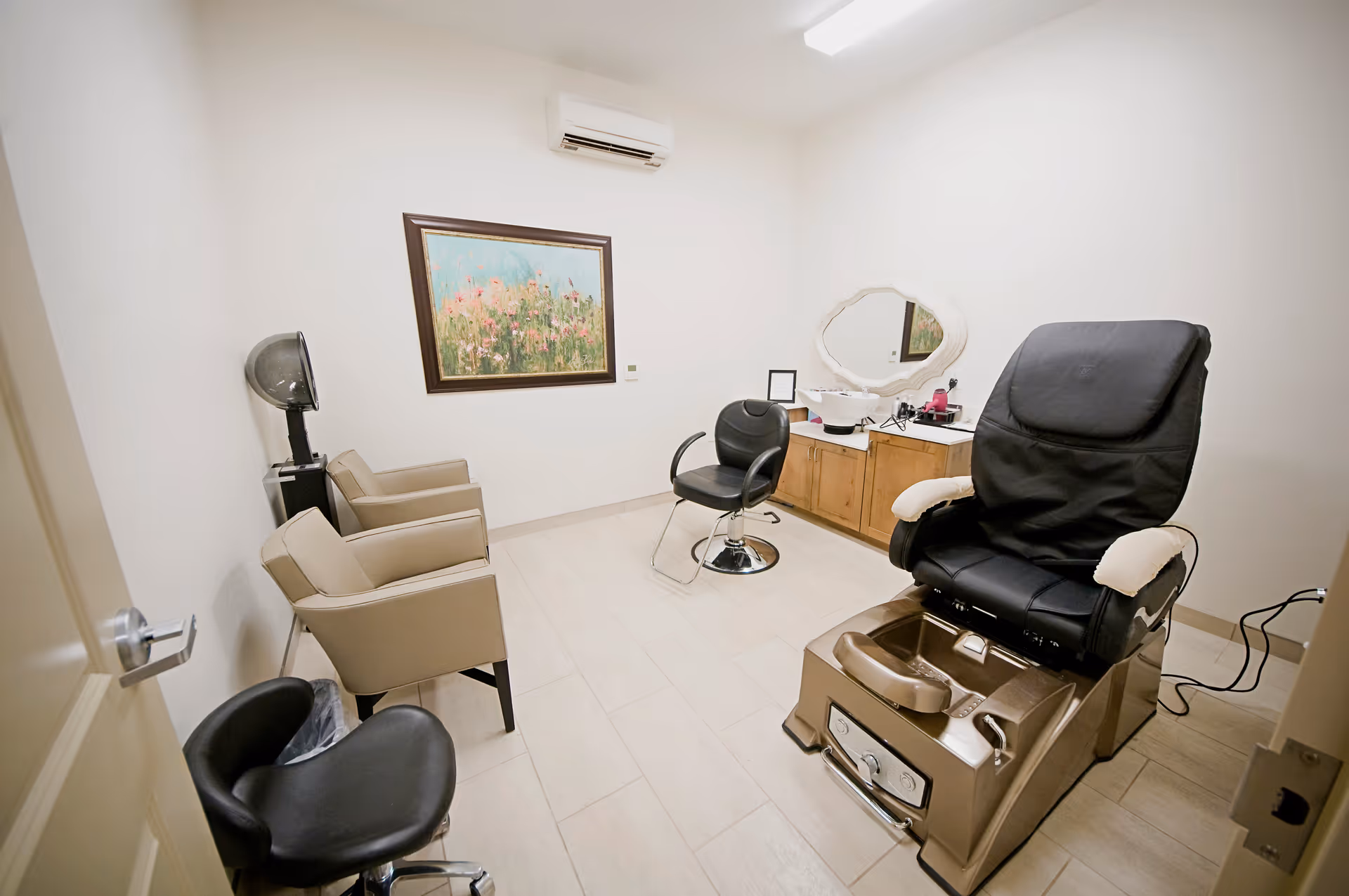 A small salon room with a pedicure chair, a black swivel chair, two beige armchairs, a hair dryer, a wooden cabinet with a mirror, and a framed floral painting on the wall.