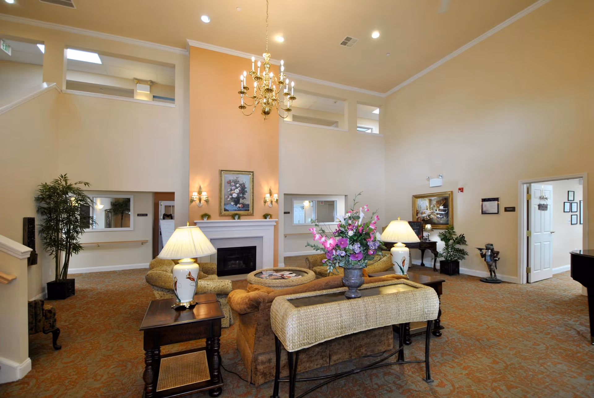 A spacious and well-lit living room area in a senior living facility with high ceilings, a chandelier, and a fireplace. The room is furnished with comfortable sofas, side tables with lamps, a floral centerpiece on a table, and decorative plants. There are paintings on the walls and an open doorway leading to another room.