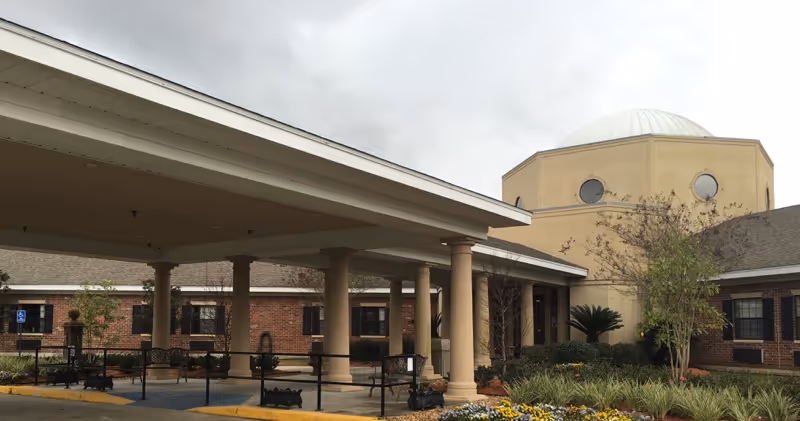 Covered front entrance (porte-cochere) of Heritage Manor Opelousas with columns, brick facade and landscaped flowerbeds.