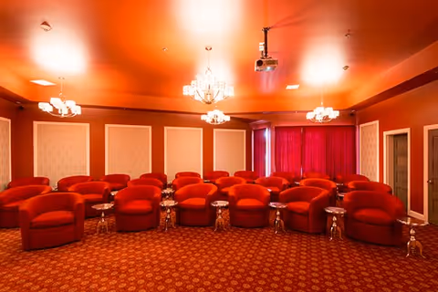A cozy theater room with multiple rows of red upholstered armchairs arranged in a semi-circle facing a projector. The room has red walls and carpet with patterned designs, chandeliers hanging from the ceiling, and red curtains covering the windows.