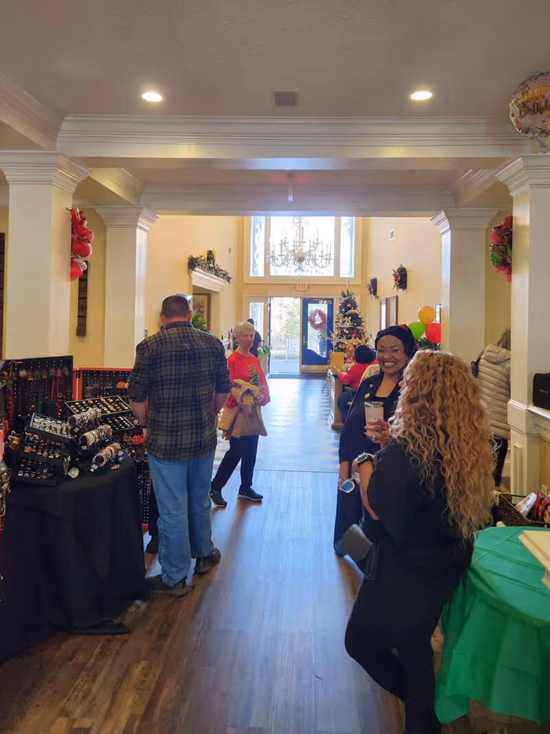 Interior hallway of Regency House Buford decorated for the holidays with a Christmas tree and wreaths. Several people are standing and conversing near tables displaying jewelry and other items. The space has wooden flooring, cream-colored walls, and a chandelier near the entrance door with glass panels.