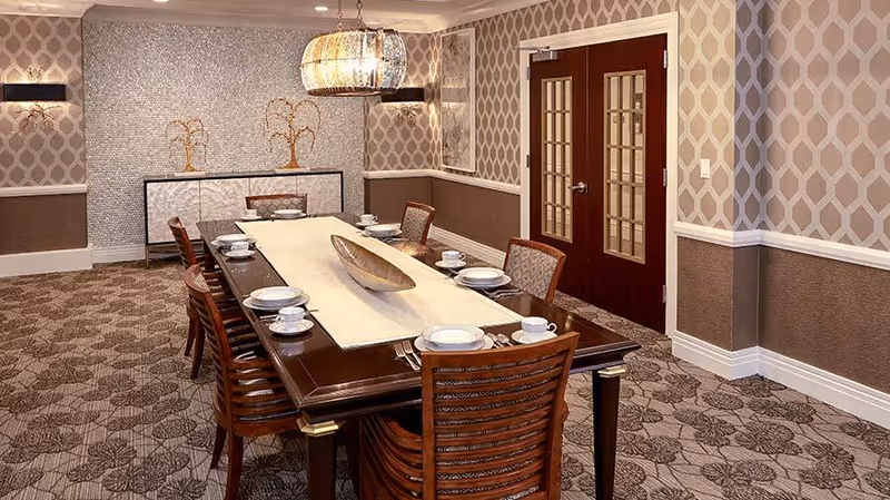 A formal dining room with a long wooden table set with plates and cups, wooden chairs, patterned wallpaper and double doors.