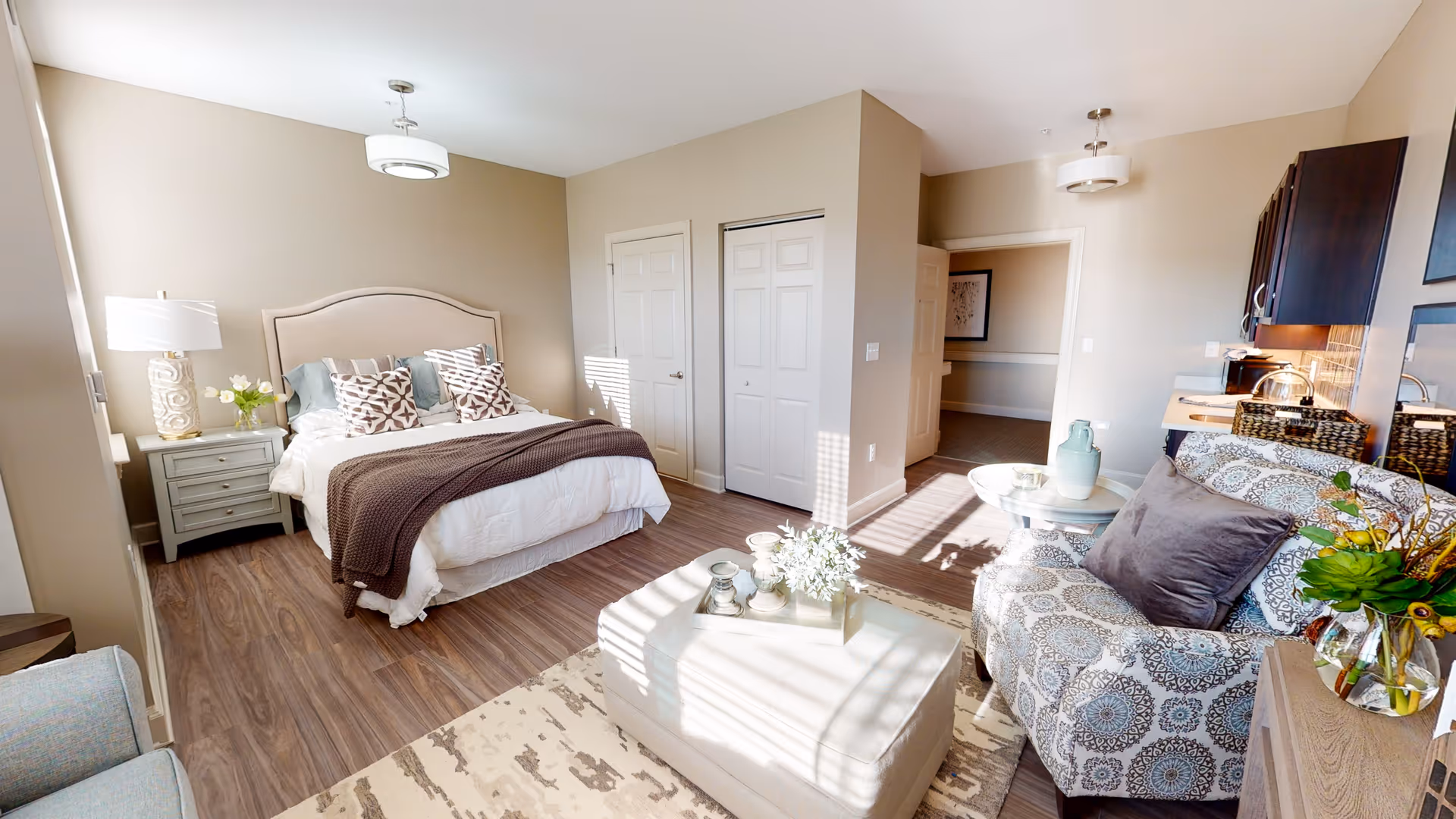 A bright and cozy bedroom with a large bed featuring multiple pillows and a brown throw blanket. There is a nightstand with a lamp and flowers on the left side of the bed. The room has wood flooring and beige walls. To the right, there is a patterned armchair with a purple pillow, a small round table with decorative items, and a cushioned ottoman in the center. The room is well-lit with natural light coming through a window, and there is a doorway leading to another room.