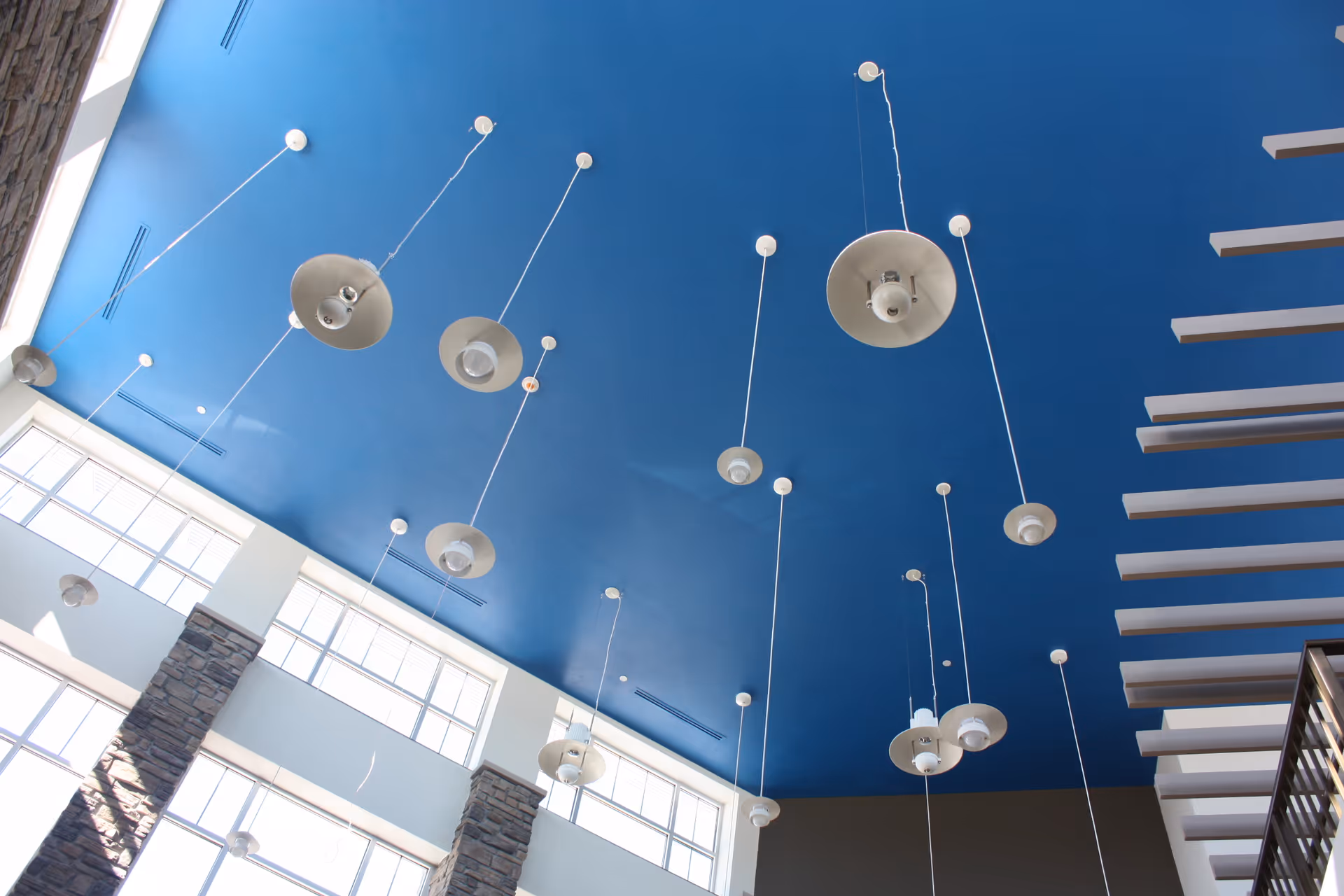 View of a high ceiling painted blue with multiple hanging pendant lights in a spacious interior area with large windows and stone pillars.
