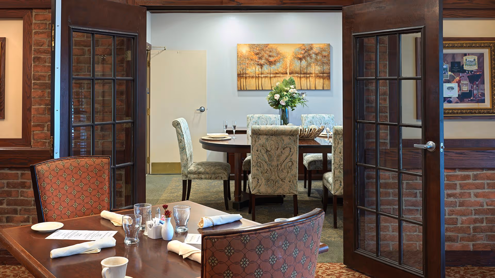 Interior view of a dining area in a senior living facility with a wooden table set with napkins, glasses, and a coffee cup. Behind it, a separate room with a round table surrounded by upholstered chairs and a vase with flowers is visible through open glass-paneled double doors. The walls have brick accents and framed artwork.