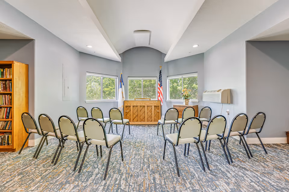 Small meeting room with chairs arranged in rows facing a wooden podium or piano beneath windows and flags.
