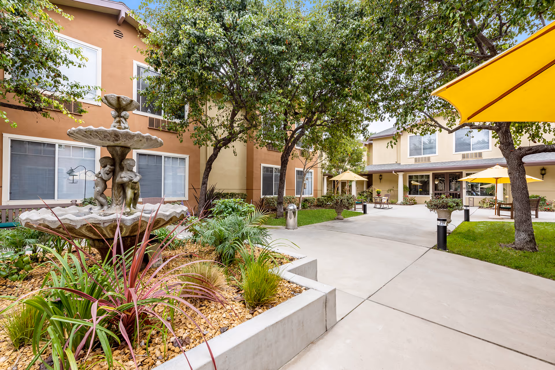 Courtyard of Cypress Place with a decorative fountain, trees, walkways and seating areas with yellow umbrellas in front of the building.