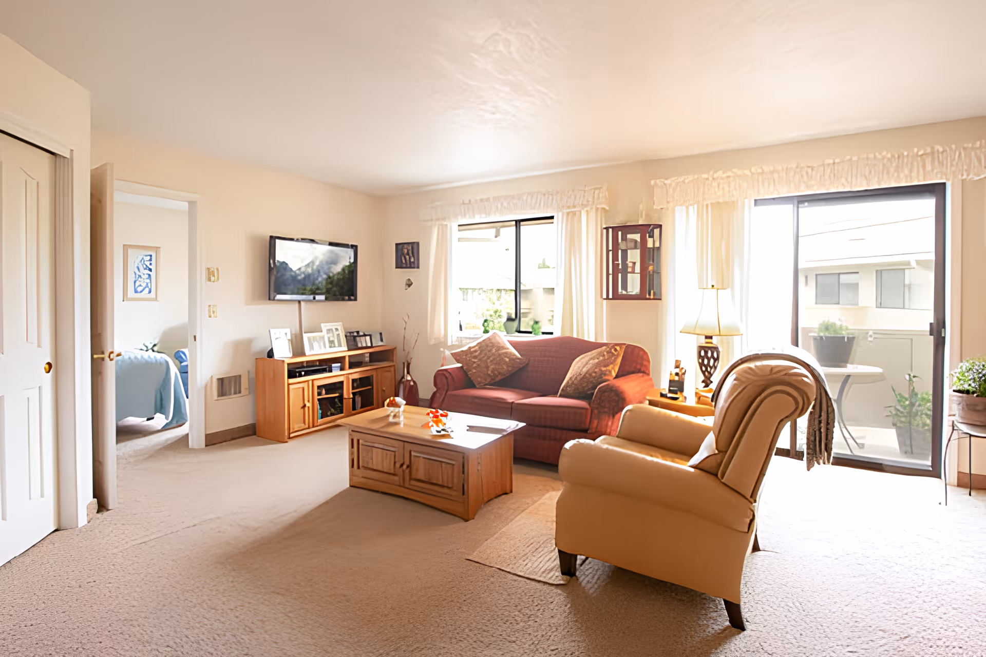 A bright living room with beige carpet and cream-colored walls. The room features a red sofa with two pillows, a beige armchair, a wooden coffee table, and a wooden TV stand with a flat-screen TV mounted above it. There are two large windows with white curtains and a sliding glass door leading to an outdoor patio. A bedroom with a blue bedspread is visible through an open door on the left.