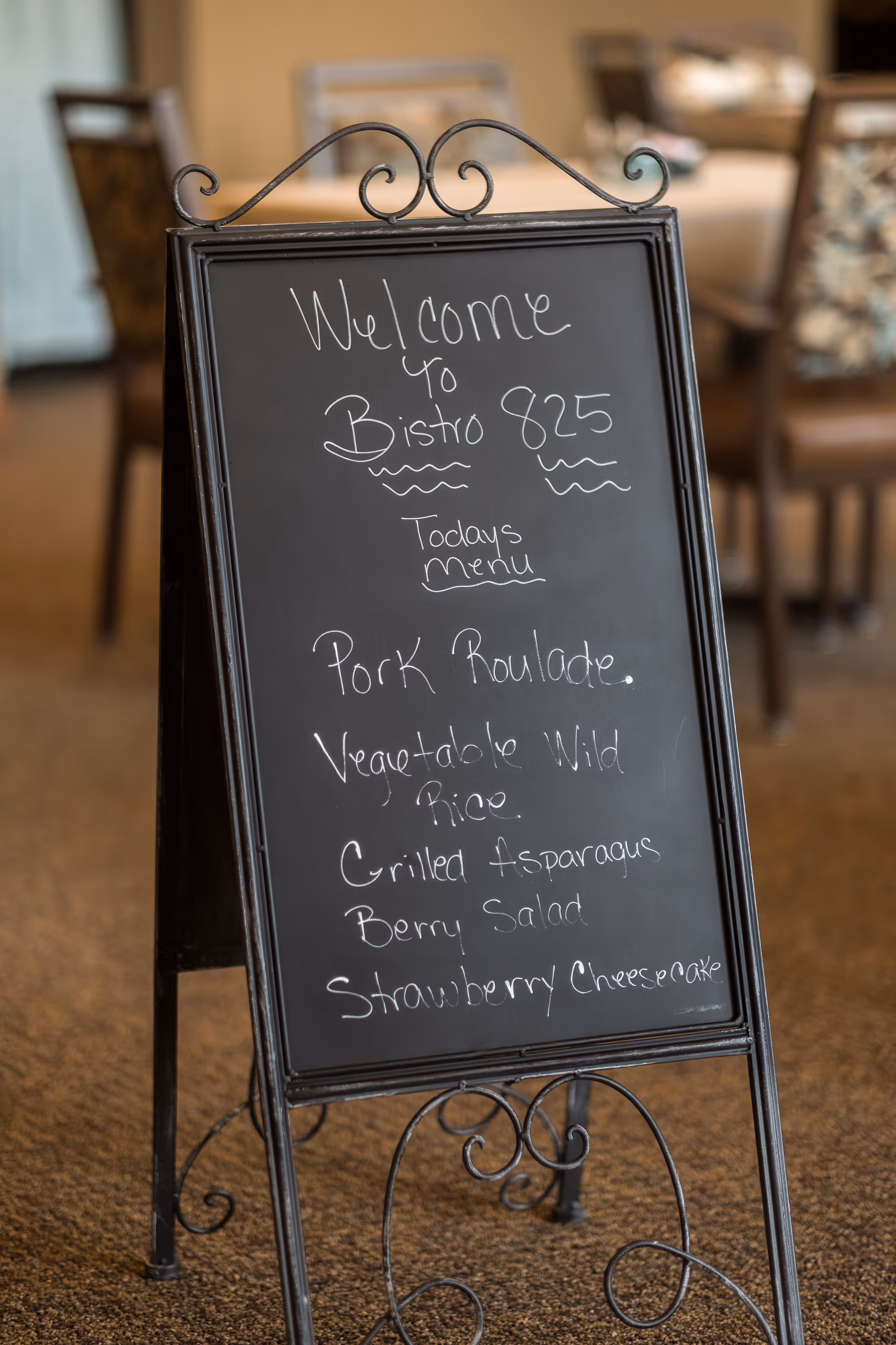 A decorative black chalkboard sign with a wrought iron frame displaying the daily menu at Bistro 825. The menu includes Pork Roulade, Vegetable Wild Rice, Grilled Asparagus, Berry Salad, and Strawberry Cheesecake. The background shows a dining area with chairs and tables.