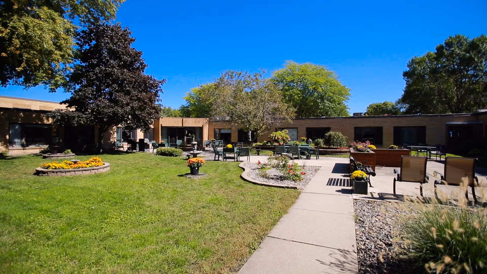 Outdoor courtyard area of a senior living community with green grass, flower beds, trees, and multiple seating areas including tables and chairs under a clear blue sky.